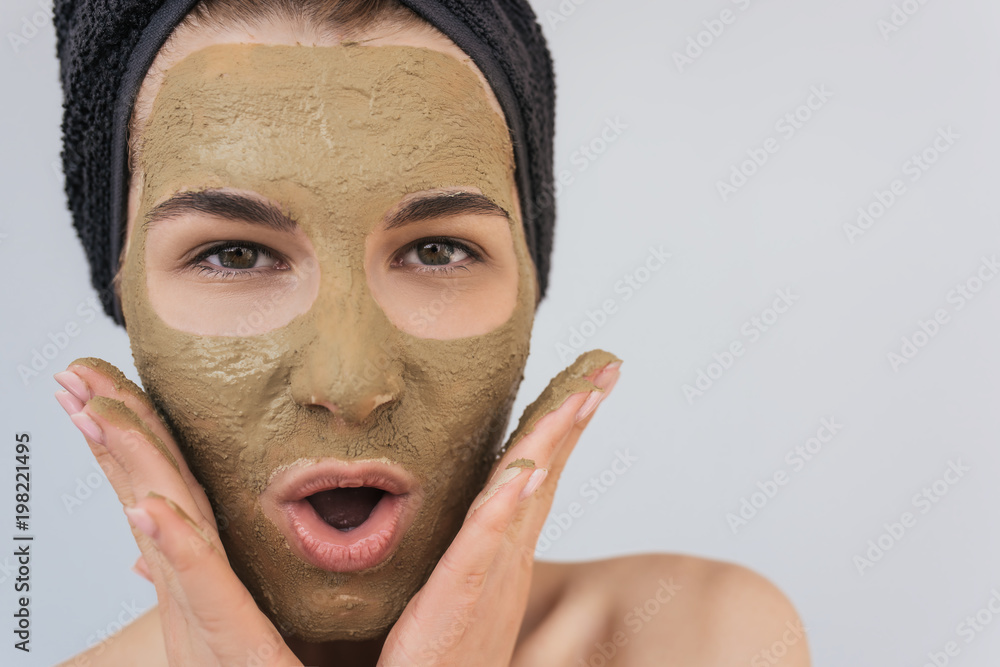 Fototapeta premium Closeup studio portrait of happy amazed young woman applying facial cosmetic clay organic mask on her face, wears black towel on hair. Female taking care of her face skin, isolated over white wall