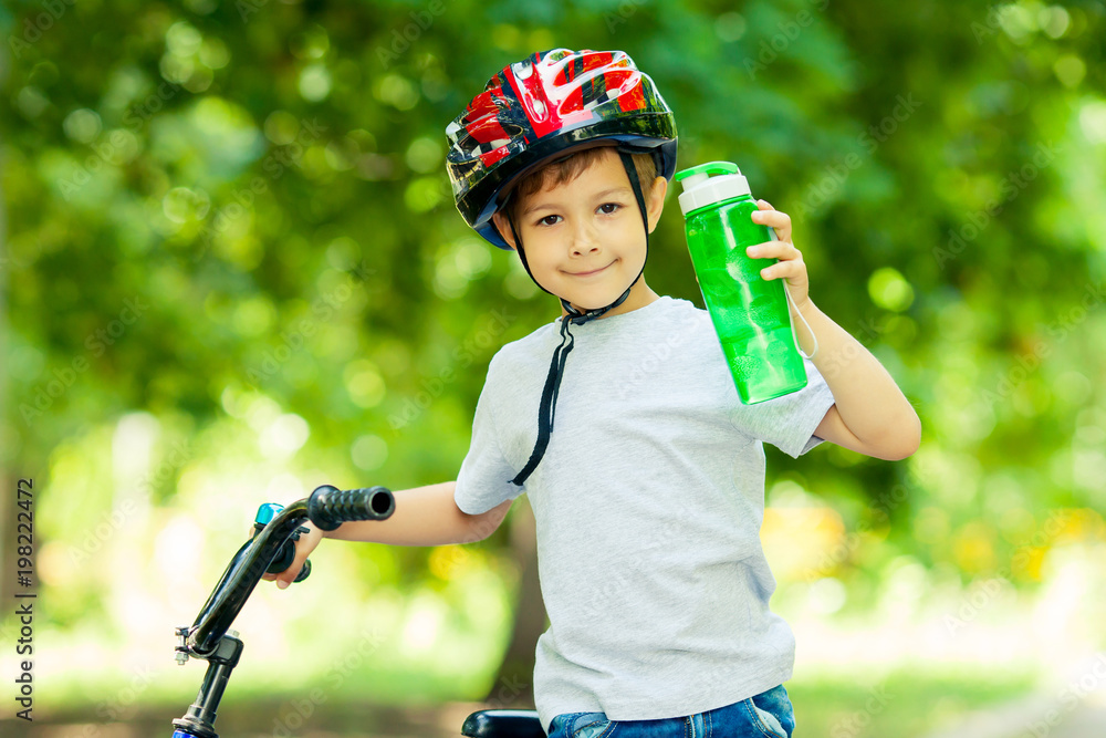 Naklejka premium Little boy drinking water by the bike. Happy smiling child in helmet riding a cycling.