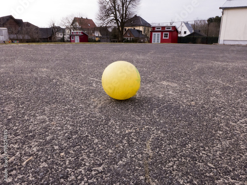 Ein gelber Ball liegt auf einem Schulhof, im Hintergrund sind zwei rote Gartenhäuser © Edda Dupree