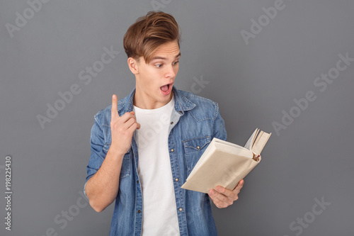 Fotografie Young man student in jeans jacket studio isolated on grey reading book pointing