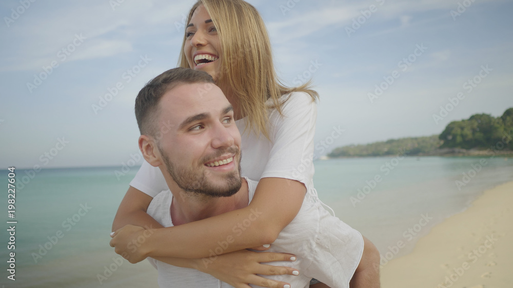 Beautiful happy couple having fun, doing piggyback on tropical sandy beach over sea and sky background Beautiful happy couple having fun, doing piggyback on tropical sandy beach over sea and sky background