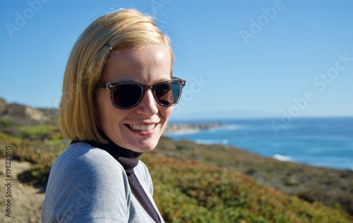 Happy blonde woman enjoying the beautiful coastal view in California (USA): Pacific ocean with limestone rock cliffs with crashing waves and a clear blue summer sky without clouds