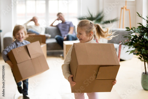 Active children enjoying moving day running carrying boxes, excited kids laughing playing in new home while parents take break to rest, happy girl and boy have fun together, family relocation concept