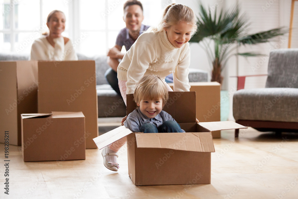 Happy siblings having fun moving into new home, smiling sister helping excited little boy riding in cardboard box, cute kids playing together in living room, family with children relocating concept Happy siblings having fun moving into new home, smiling sister helping excited little boy riding in cardboard box, cute kids playing together in living room, family with children relocating concept