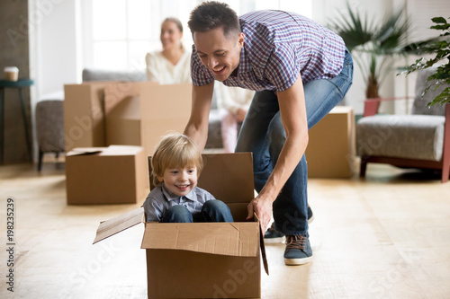 Father helping cute little son riding in box, happy dad playing with small boy after relocation, young family kids having fun in new house living room, excited child and daddy on moving day concept