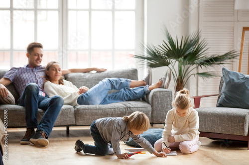 Children sister and brother playing drawing together on floor while young parents relaxing at home on sofa, little boy girl having fun, friendship between siblings, family leisure time in living room photo