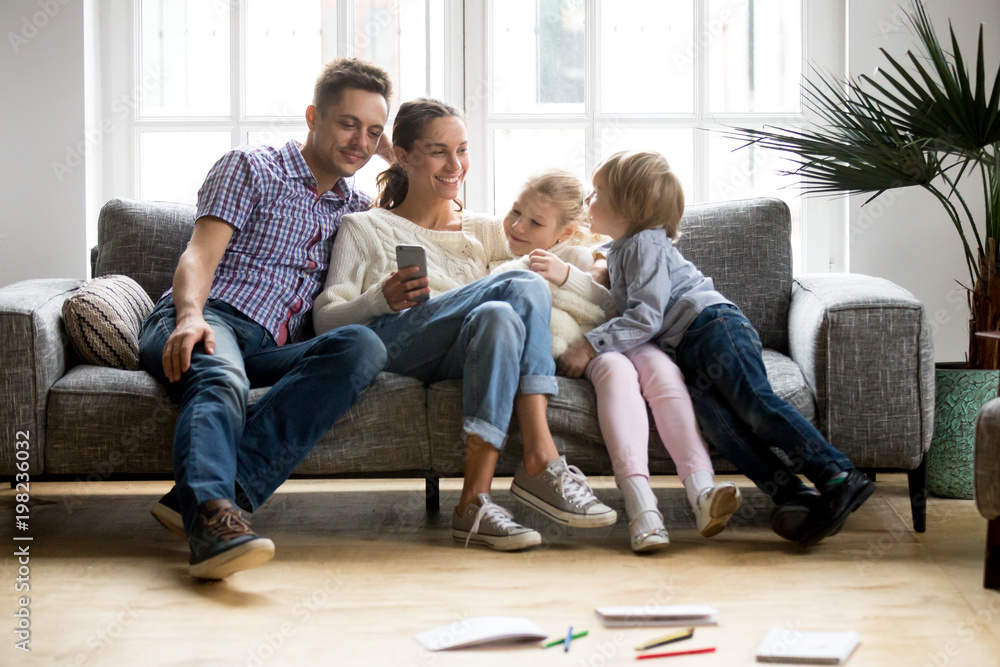 Happy family with children shopping with mobile app together, smiling young couple buying online using smartphone sitting on sofa with kids, parents and son daughter entertaining with phone at home Happy family with children shopping with mobile app together, smiling young couple buying online using smartphone sitting on sofa with kids, parents and son daughter entertaining with phone at home
