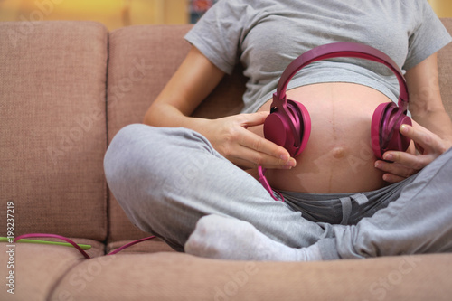 Pregnant woman sitting on a couch and holding headphones on belly. Unborn child listens to music with headphones.