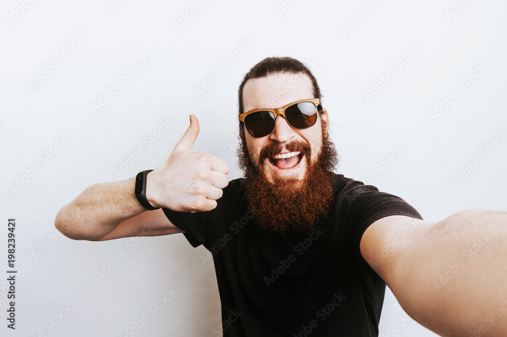 Naklejka premium Cheerful bearded man wearing sunglasses and showing thumbs up gesture over white background