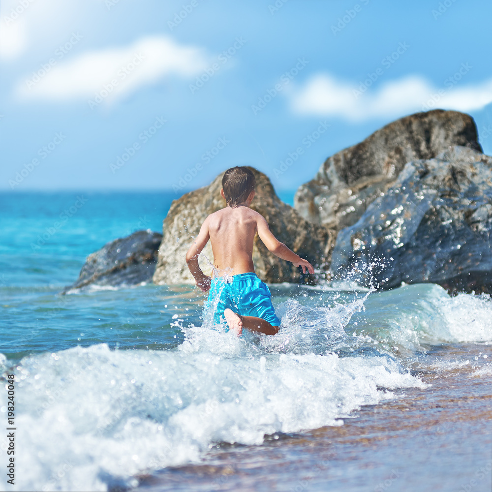 Fototapeta premium Cute Caucasian boy is running in the water along the sea shore. Back view.
