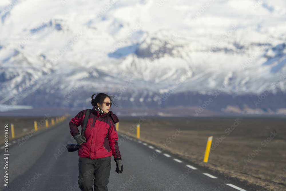 Portrait of asian traveler photographer lady in red warm cloth walking on road with snow capped mountain in background,travel in winter Iceland theme. Portrait of asian traveler photographer lady in red warm cloth walking on road with snow capped mountain in background,travel in winter Iceland theme.