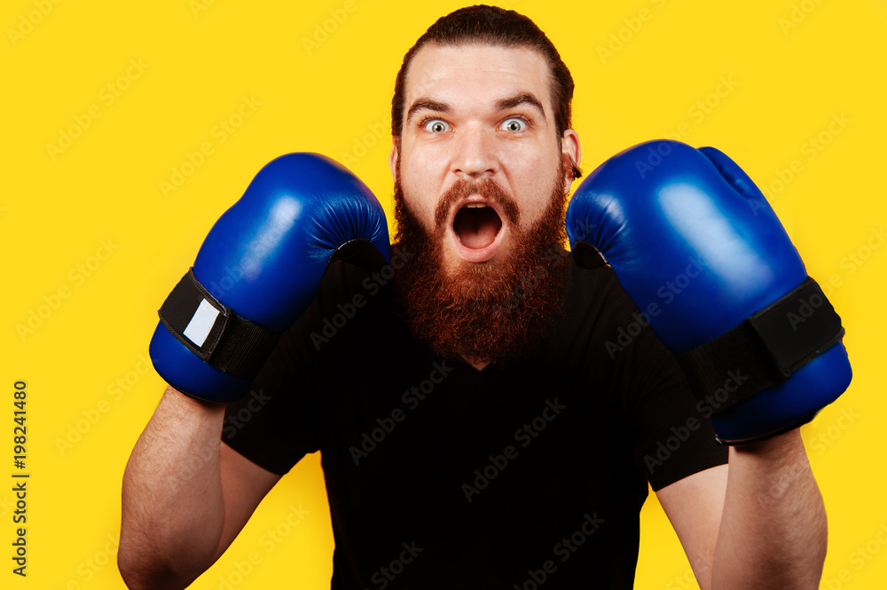 Portrait of a shocked bearded man with blue boxing gloves isolated on yellow background Portrait of a shocked bearded man with blue boxing gloves isolated on yellow background