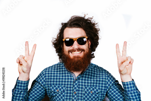 Portrait of a young man with beard, brunet, curly haired man gesturing with two fingers near eyes on a white background. Peace love concept. Smiling happy bearded hipster. Happy man with sunglasses.