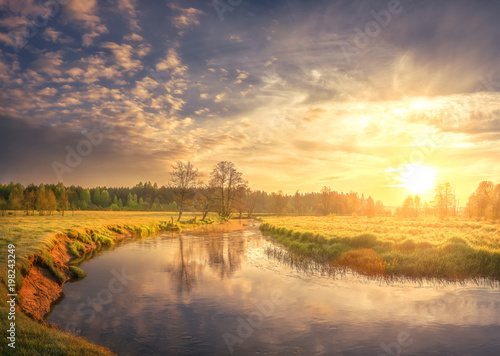 Nature landscape of spring river on morning dawn. Bright sun lights on green grass and young foliage. Rural scene. Trees on shore of river and colorful sky with clouds shining on warm sunlight.