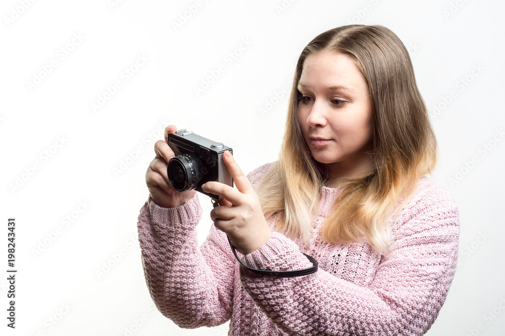 Portrait of a young beautiful happy girl wearing pale pink knitted sweater looking at a vintage camera in her hands against white background. Lifestyle and people concept Portrait of a young beautiful happy girl wearing pale pink knitted sweater looking at a vintage camera in her hands against white background. Lifestyle and people concept