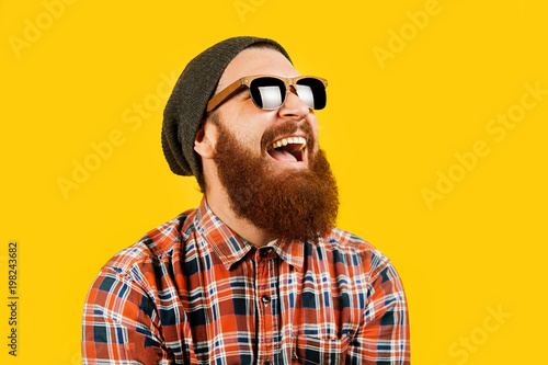 Portrait of young hipster man in sunglasses and hat posing on yellow background. Smiling bearded man wearing sunglasses, studio shot. Happy man with beard looking up.