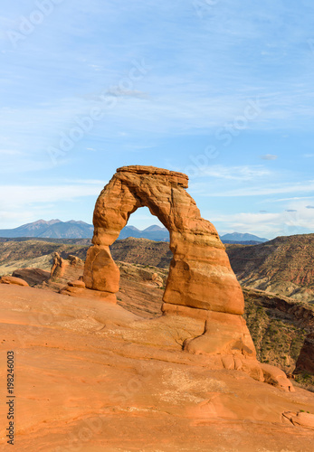 Sunset at Delicate Arch - Vertical - Colorful Delicate Arch in bright evening sun, with rolling rocky mountains in the background, Arches National Park, Moab, Utah, USA.