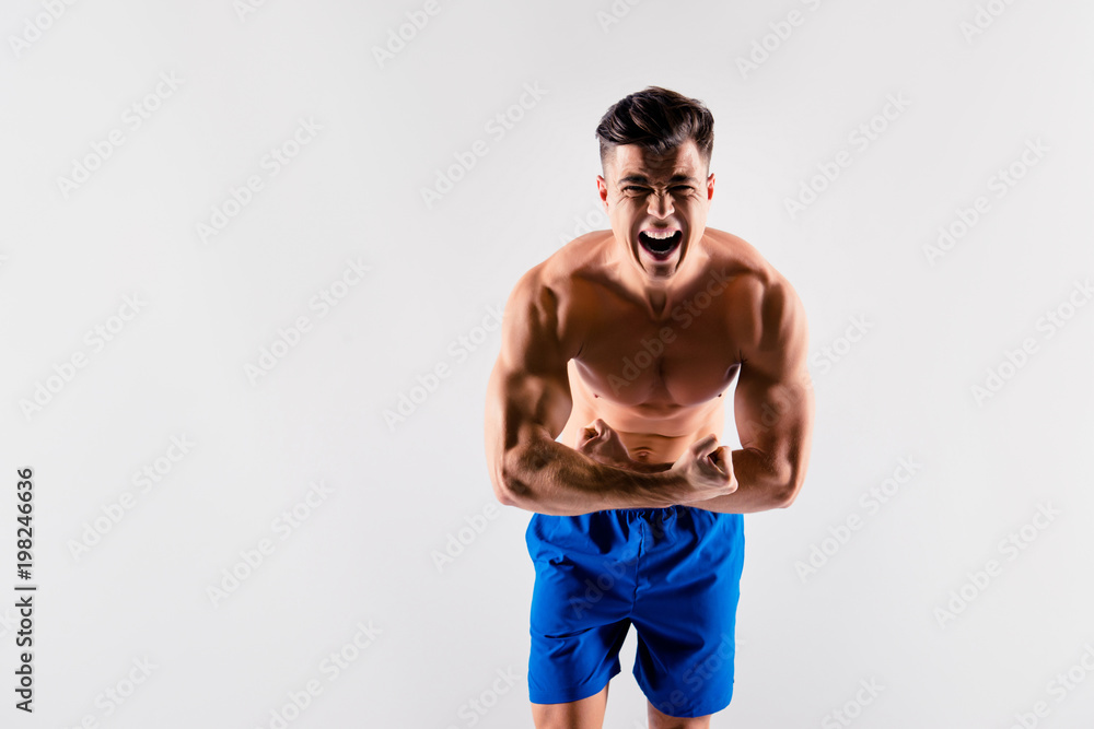Portrait of aggressive screaming handsome athlete wearing blue loose shorts, he is demonstrating and toughening his muscular body, isolated on grey background Portrait of aggressive screaming handsome athlete wearing blue loose shorts, he is demonstrating and toughening his muscular body, isolated on grey background
