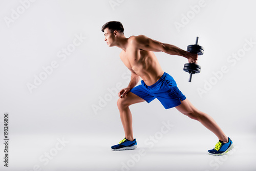 Side profile view photo of handsome virile confident purposeful shirtless muscular guy leaning on the leg and lifting a dumbbell from behind his back  Isolated on grey background  copy-space