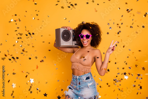 Hey what's up! Join the get-together! Cool carefree attractive reckless crazy female party-goer is holding a stereo boombox on a shoulder and showing v-sign, isolated on yellow background