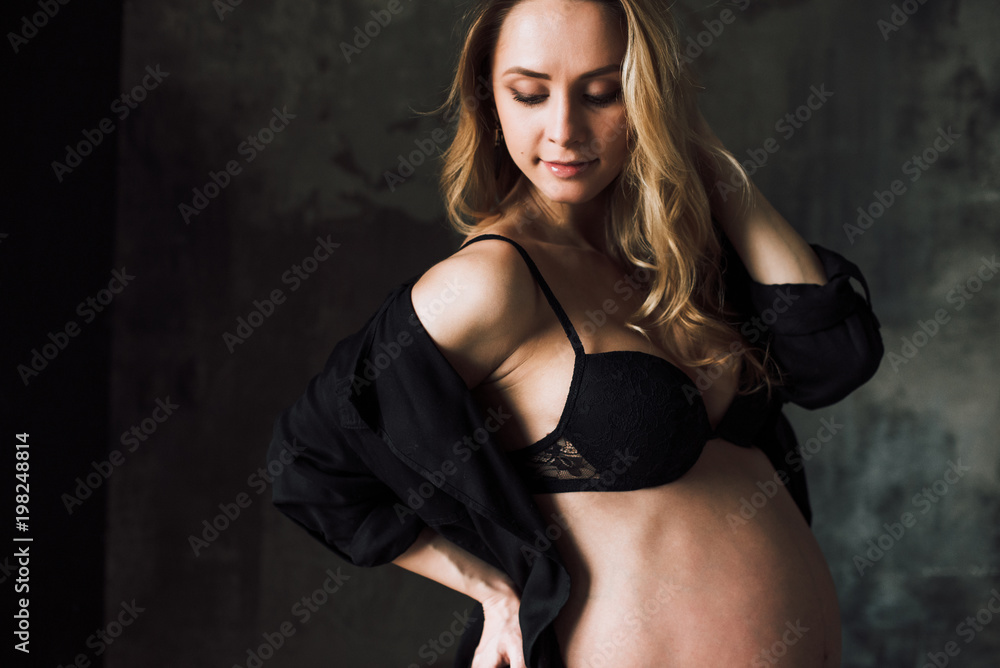 Pregnant woman. Portrait of a beautiful pregnant woman in black lingerie who stands on the background of a dark wall and looks down Pregnant woman. Portrait of a beautiful pregnant woman in black lingerie who stands on the background of a dark wall and looks down