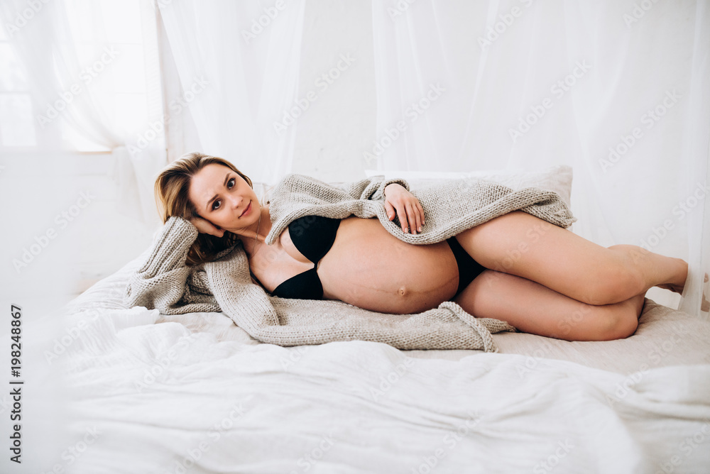 Naklejka premium Pregnant woman. Beautiful blonde pregnant woman in black lingerie with charming smile lies on the bed in the bedroom