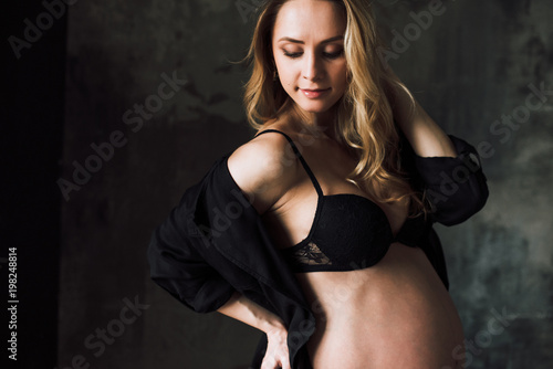 Pregnant woman. Portrait of a beautiful pregnant woman in black lingerie who stands on the background of a dark wall and looks down