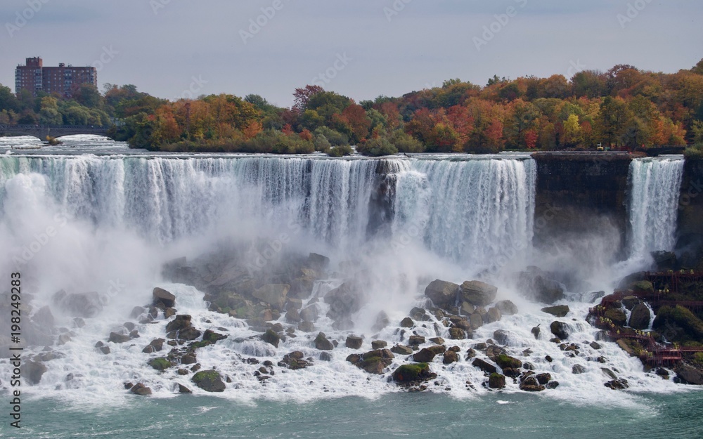 Fototapeta premium Beautiful and impressive panorama of the Niagara Falls in Ontario (Canada) on a bright autumn day with water crashing down the falls onto rocks creating lots of mist in front of the city skyline