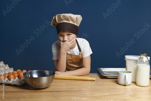 Portrait of frustrated 10 year old boy in chef uniform covering mouth feeling puzzled while going to make pancakes by himself for the first time with milk, eggs, flour and utensils on wooden table photo