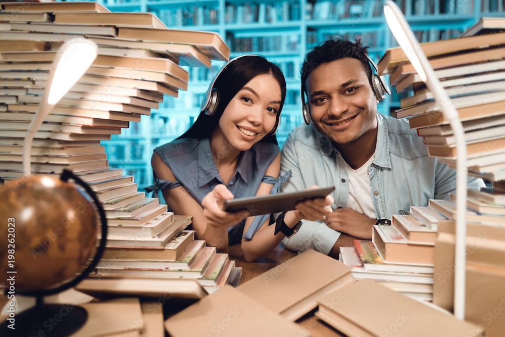 Ethnic asian girl and indian mixed race guy surrounded by books in library. Students are using tablet. Ethnic asian girl and indian mixed race guy surrounded by books in library. Students are using tablet.