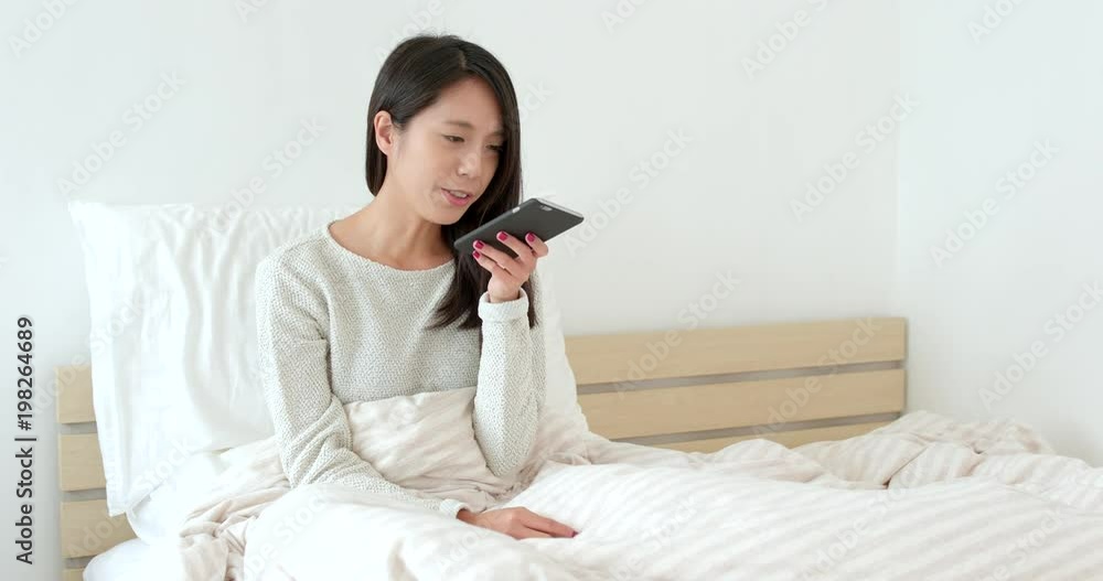 Woman sending audio message with smart phone and sitting on bed Woman sending audio message with smart phone and sitting on bed