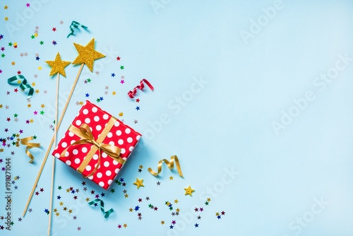 Top view of a red dotted gift box, golden magic wands, colorful confetti and ribbons over blue background. Celebration concept. Copy space.