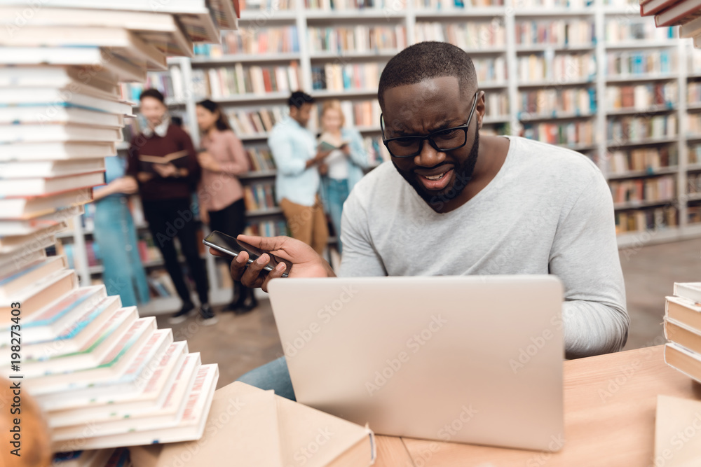 Ethnic african american guy surrounded by books in library. Student is using laptop. Ethnic african american guy surrounded by books in library. Student is using laptop.