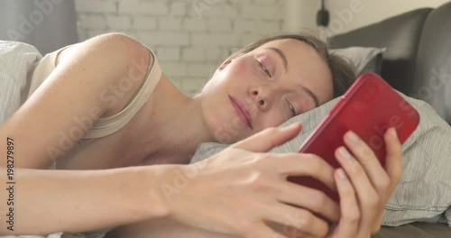 technology, internet, communication and people concept - happy smiling young woman texting on smartphone in bed at home bedroom at nmorning photo