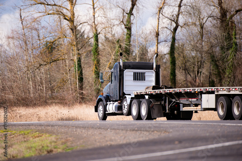 Canvas Print Big rig black semi truck with flat bed semi trailer running on winding road with