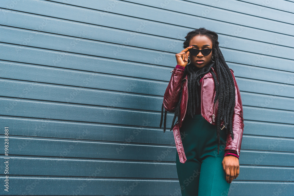 Naklejka premium portrait of beautiful stylish young african american woman adjusting sunglasses and looking at camera