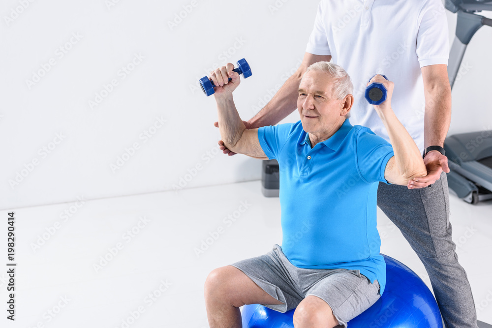 partial view of rehabilitation therapist assisting senior man exercising with dumbbells on fitness ball on grey backdrop partial view of rehabilitation therapist assisting senior man exercising with dumbbells on fitness ball on grey backdrop