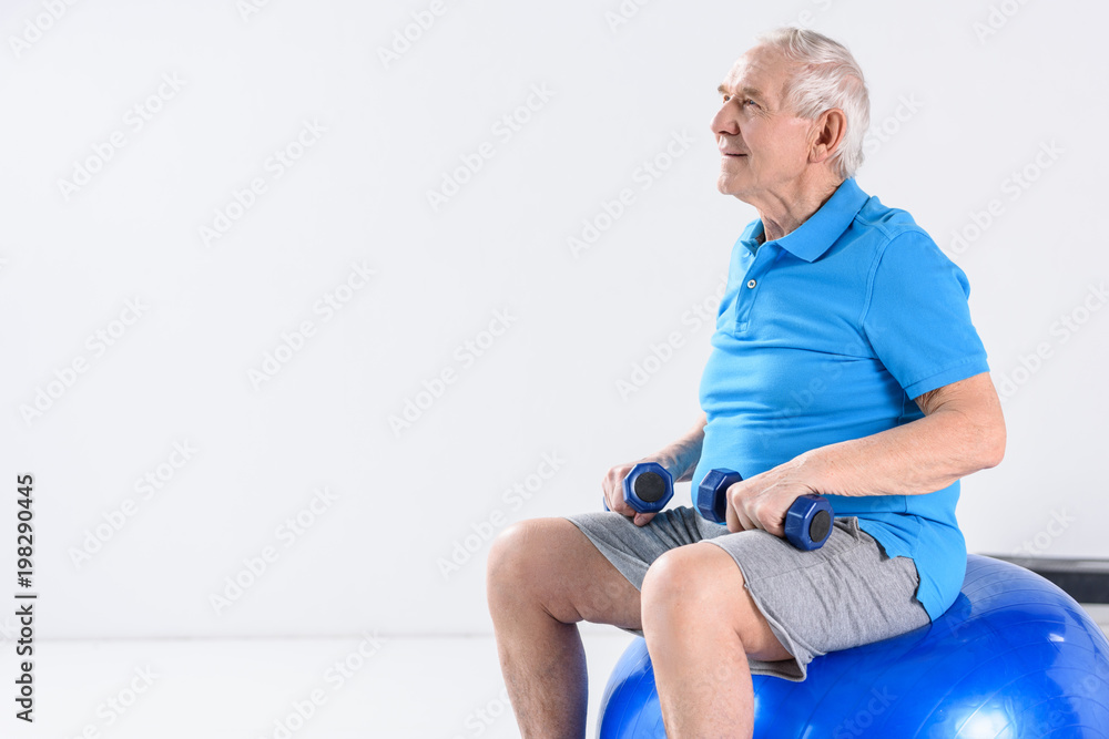 senior man with dumbbells sitting on fitness ball on grey backdrop senior man with dumbbells sitting on fitness ball on grey backdrop