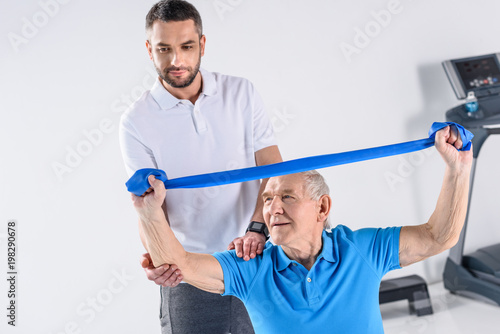 rehabilitation therapist assisting senior man exercising with rubber tape