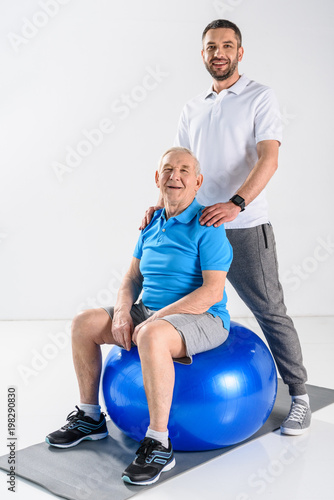 smiling rehabilitation therapist and senior man on fitness ball looking at camera on grey backdrop