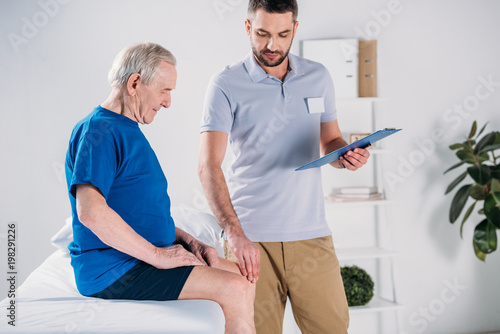 rehabilitation therapist with notepad checking senior mans knee on massage table