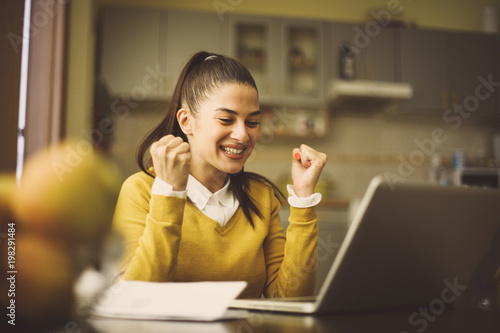 Happy young woman sitting at home and working on laptop and receiving good news. photo