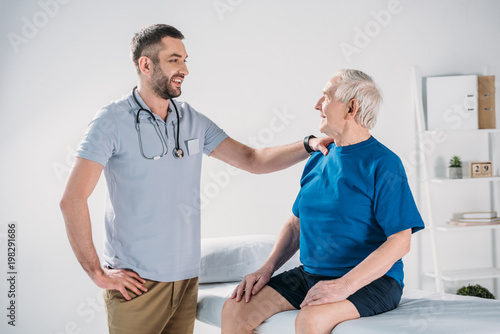 rehabilitation therapist with stethoscope and smiling senior man on massage table