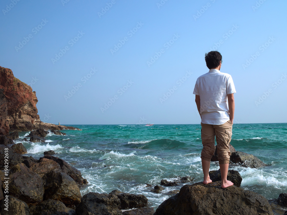 Back view of lonely Asian man standing on the stone of sea shore and looking at far away.