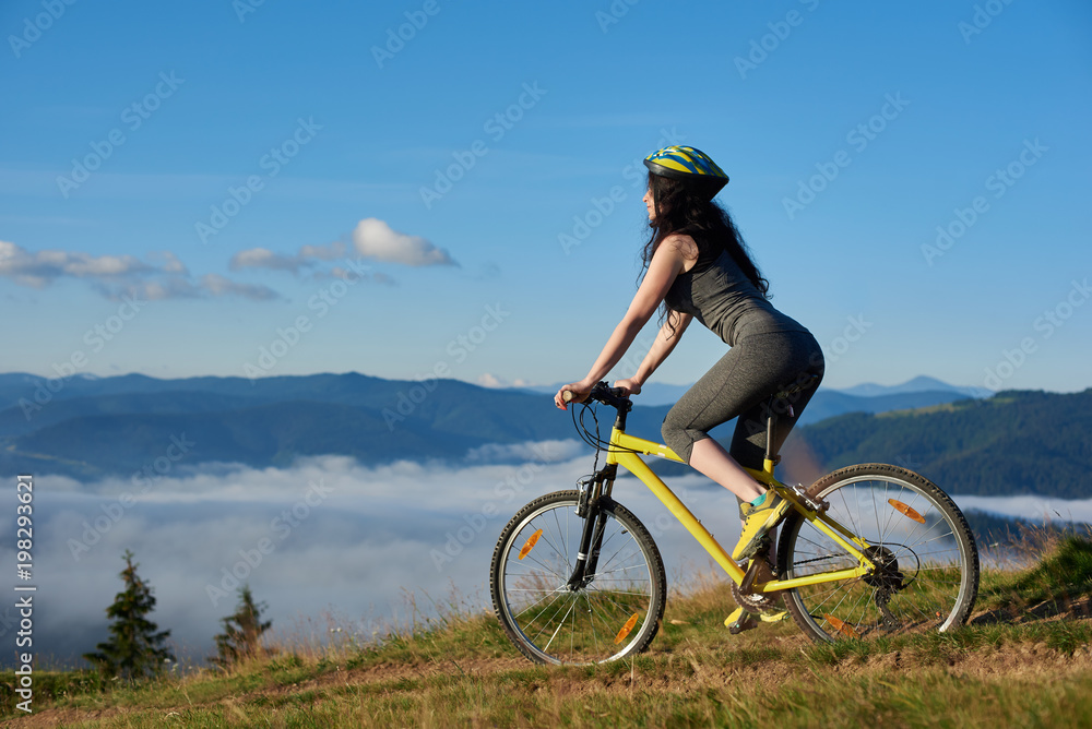 Back view of sporty girl cycling on yellow bike on rural trail in the mountains, wearing helmet, on sunny morning. Foggy mountains, forests on blurred background. Outdoor sport activity. Copy space