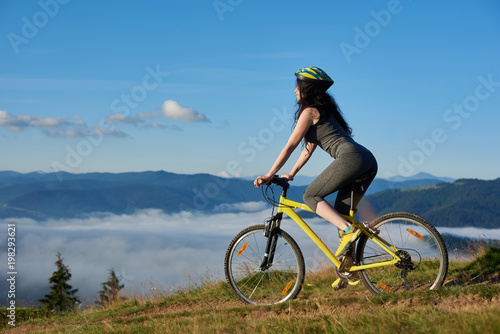 Back view of sporty girl cycling on yellow bike on rural trail in the mountains, wearing helmet, on sunny morning. Foggy mountains, forests on blurred background. Outdoor sport activity. Copy space