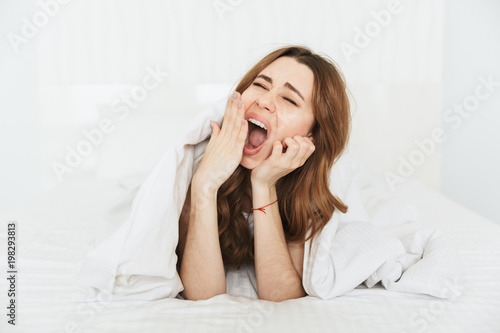 Sleepy young lady lies in bed indoors at home yawning