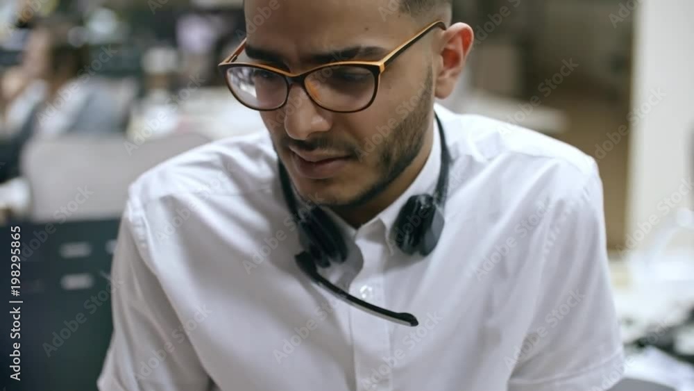 PAN of Arab male call centre agent in glasses and with headset around neck chatting with unrecognizable colleague during break PAN of Arab male call centre agent in glasses and with headset around neck chatting with unrecognizable colleague during break