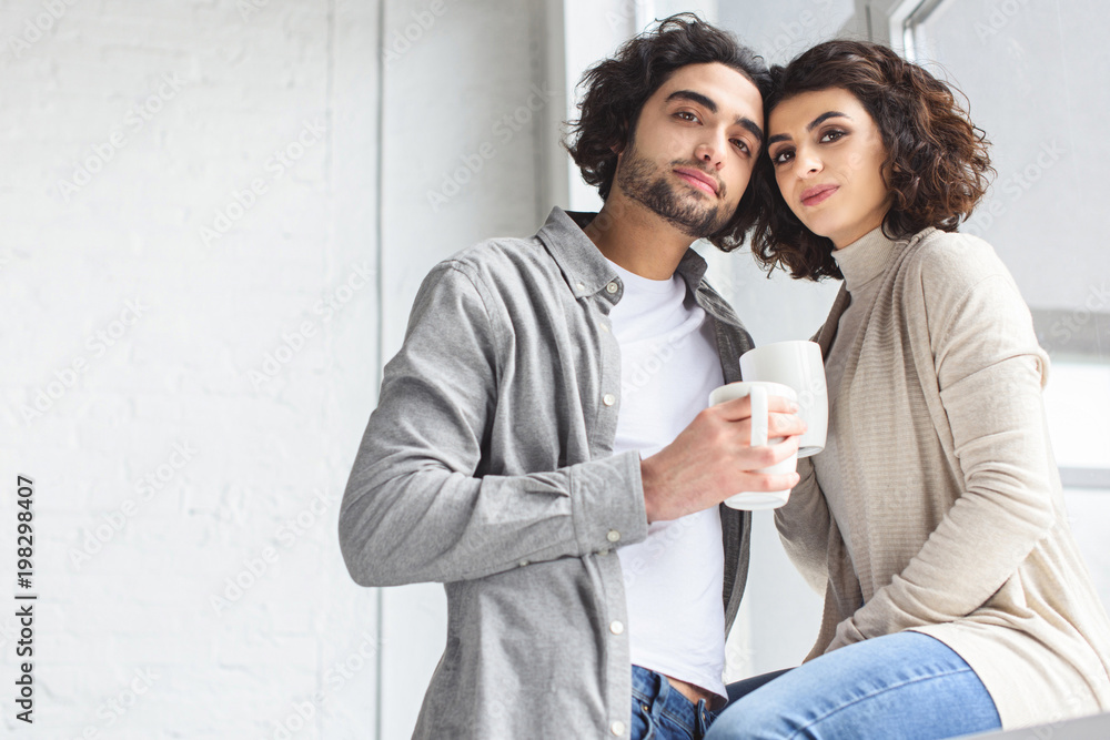young couple holding cups of coffee and looking at camera at home young couple holding cups of coffee and looking at camera at home