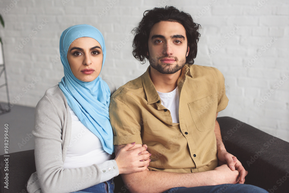 muslim couple sitting on sofa in living room and looking at camera muslim couple sitting on sofa in living room and looking at camera